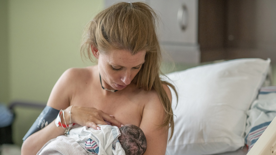 A woman is on a hospital bed holding her baby in her arms and trying to help her baby breastfeed.