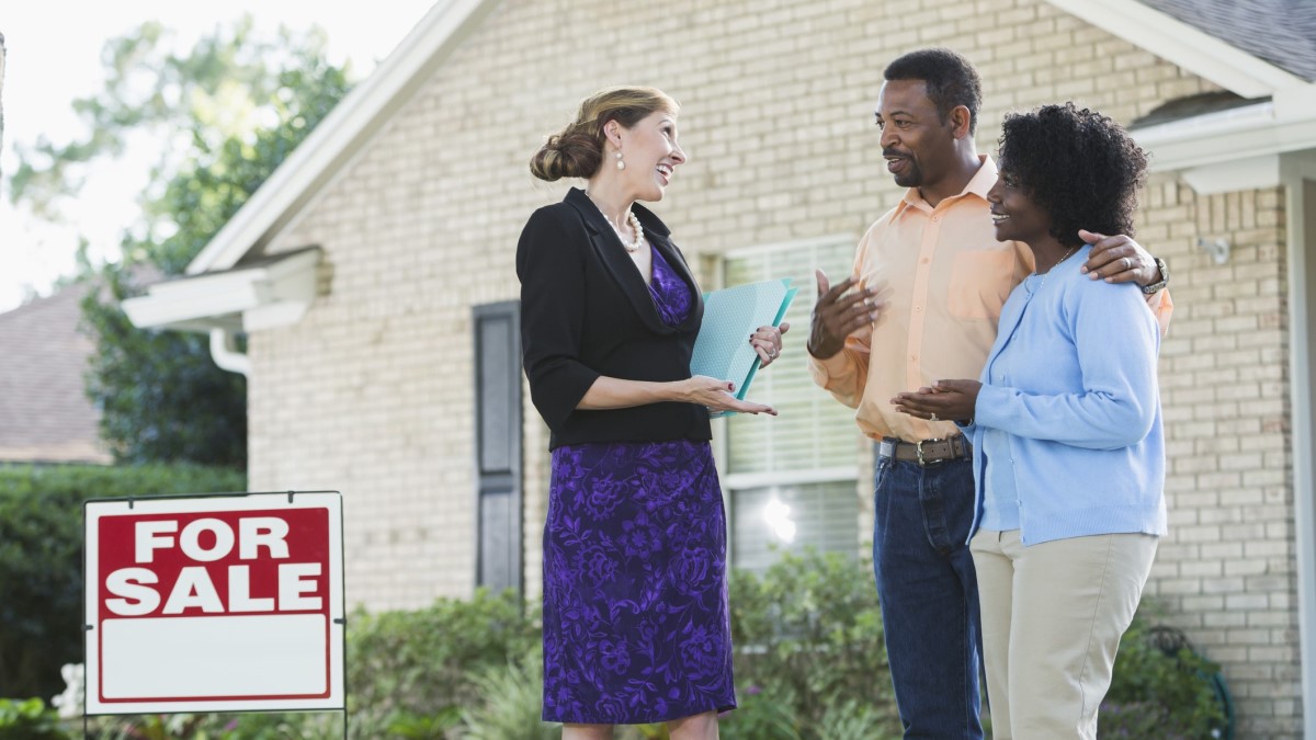 Real estate A real estate agent standing in front of a house with a For Sale sign in the yard, talking with a couple.