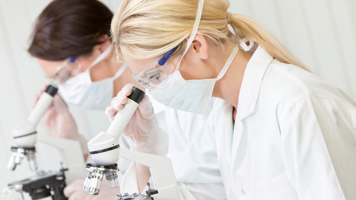 Dos técnicos de laboratorio con batas blancas, mascarillas y gafas de protección mirando a través de microscopios.