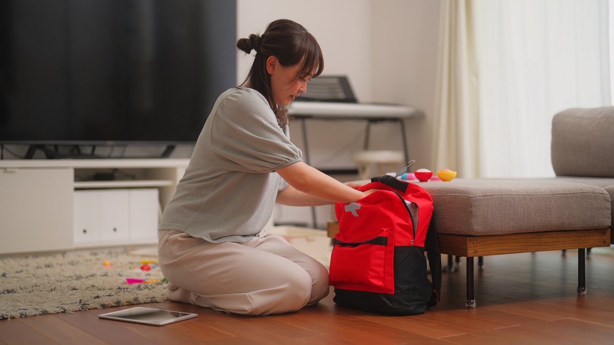 Woman packing bag A woman kneels on the floor while packing a red and black backpack, with toys scattered around and a tablet on the floor beside her.