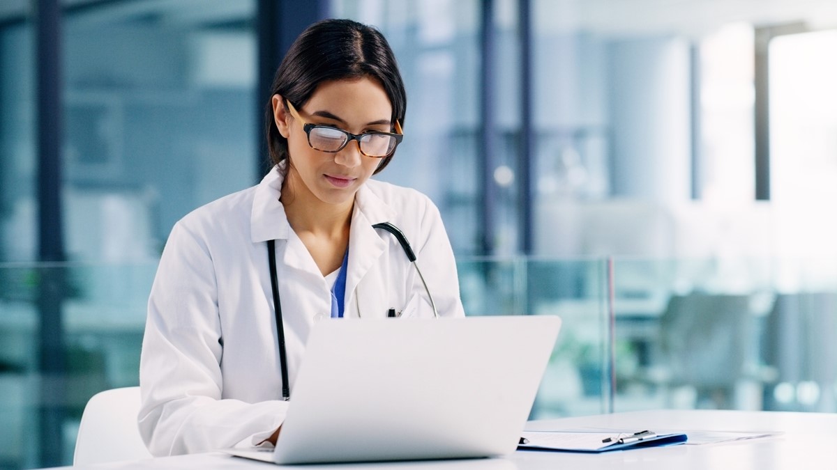 A female healthcare provider wearing glasses sits at a desk with an open notebook computer.