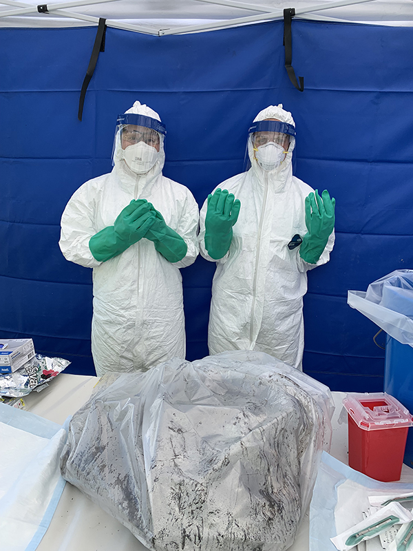Two people in white biohazard suits stand behind a table. On the table is a multi-gallon plastic bag with unknown material inside.