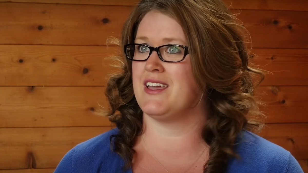 Woman wearing glasses and blue shirt with long brown hair speaking with wood-walled background.