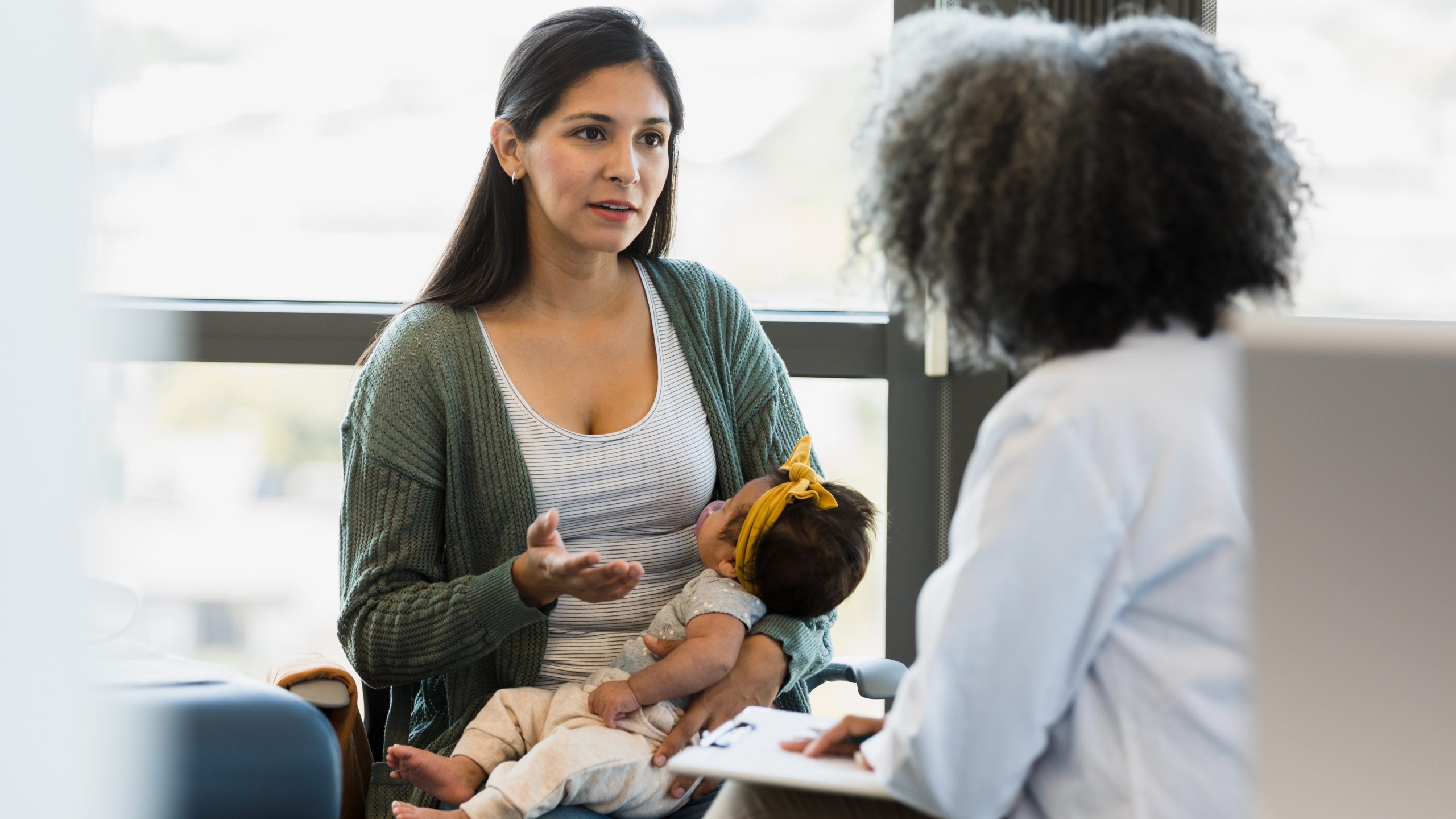 Mujer sosteniendo a una bebé y hablando con una proveedora de atención médica en el consultorio médico.