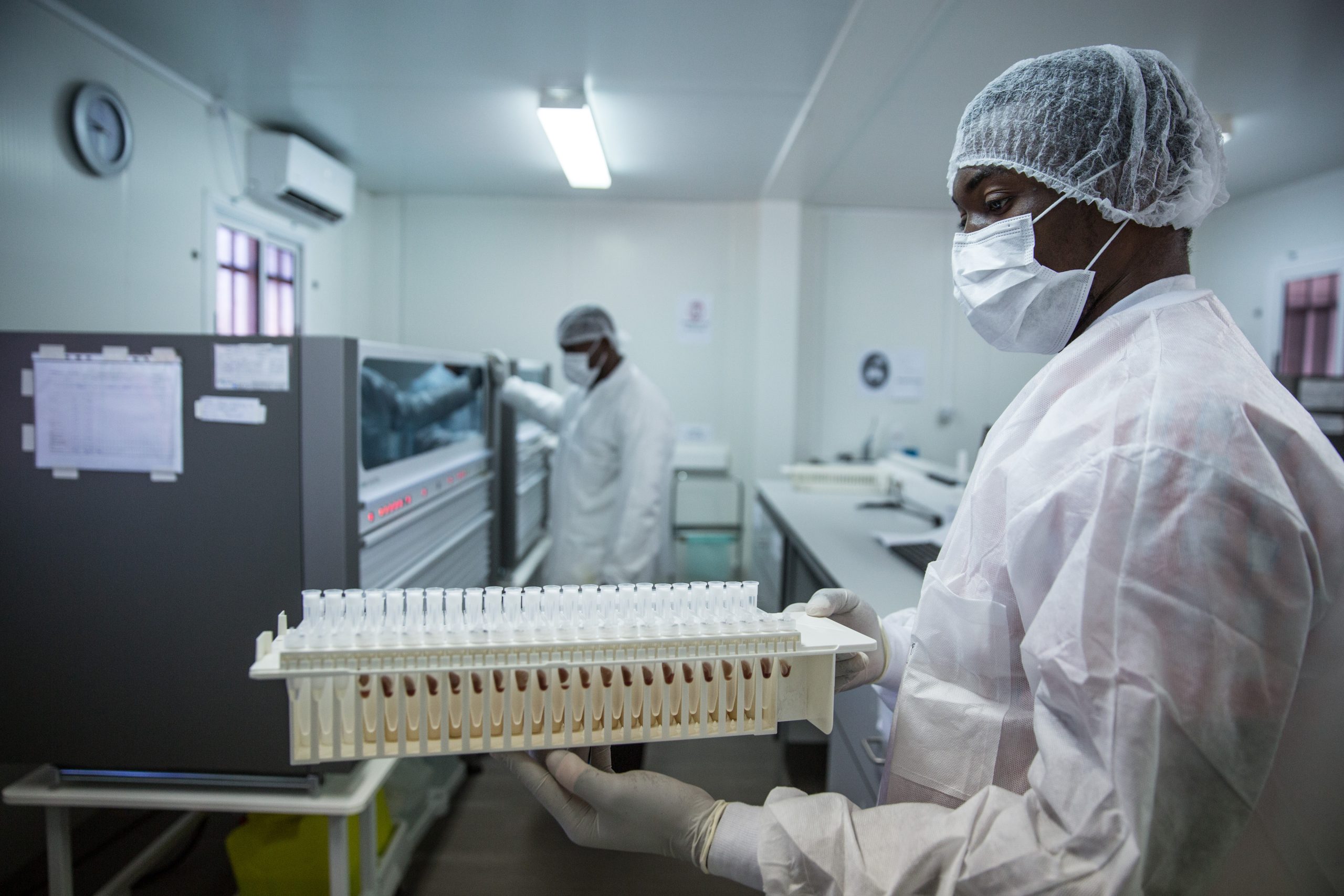 Uganda Ebola Lab Worker A Black man wearing white lab gear, blue gloves, a mask, and hair protection stands in a laboratory.
