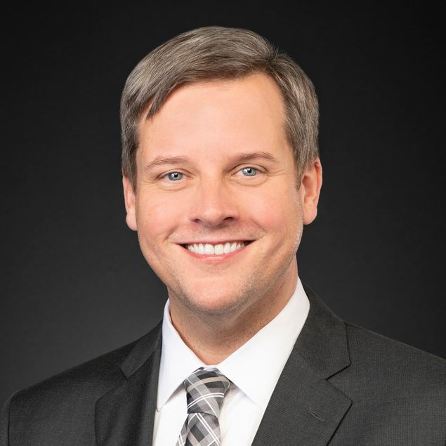 A man with gray hair against a black background. He is wearing a grey suit jacket, white shirt, and checkered tie.