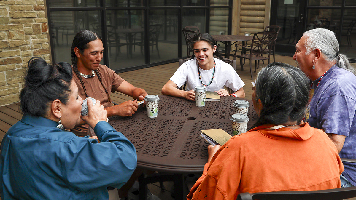 Five tribal adults sit around an outdoor table with coffee cups and books