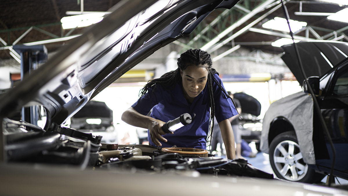-- title missing -- Una trabajadora de estaciones de servicio de automóviles inspecciona un compartimento de un motor con una luz.