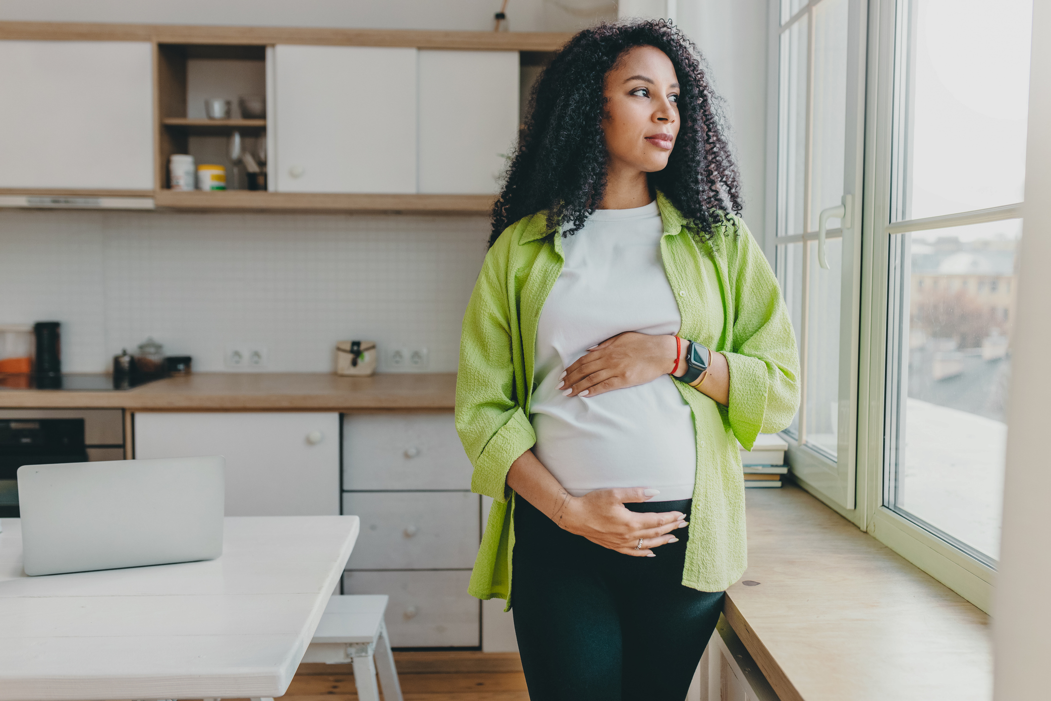 Pregnant woman of color standing and looking out the kitchen window, while holdingbelly.