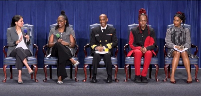 Five persons sit in chairs on a stage