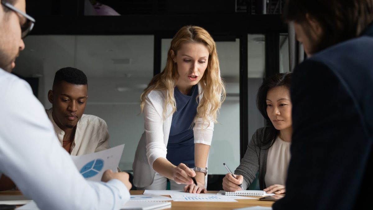 woman explaining graphs to coworkers 1200x675px A woman explaining graphs to four coworkers in a meeting room.
