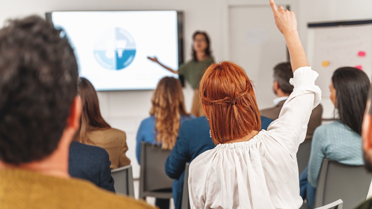 Woman raising hand during presentation 1200x675px A woman raising her hand during a presentation.