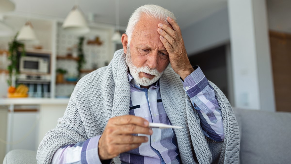An older man with a headache views a thermometer in his hand.