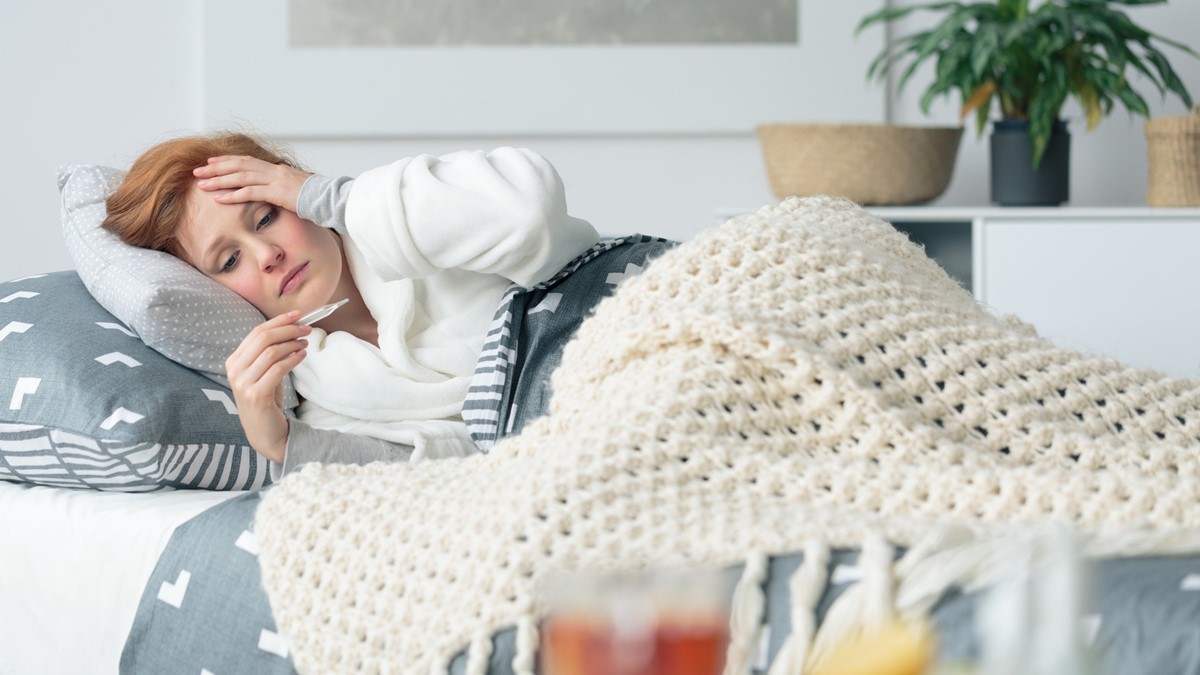 A woman feeling ill lies on a couch looking at a thermometer and holding her head.
